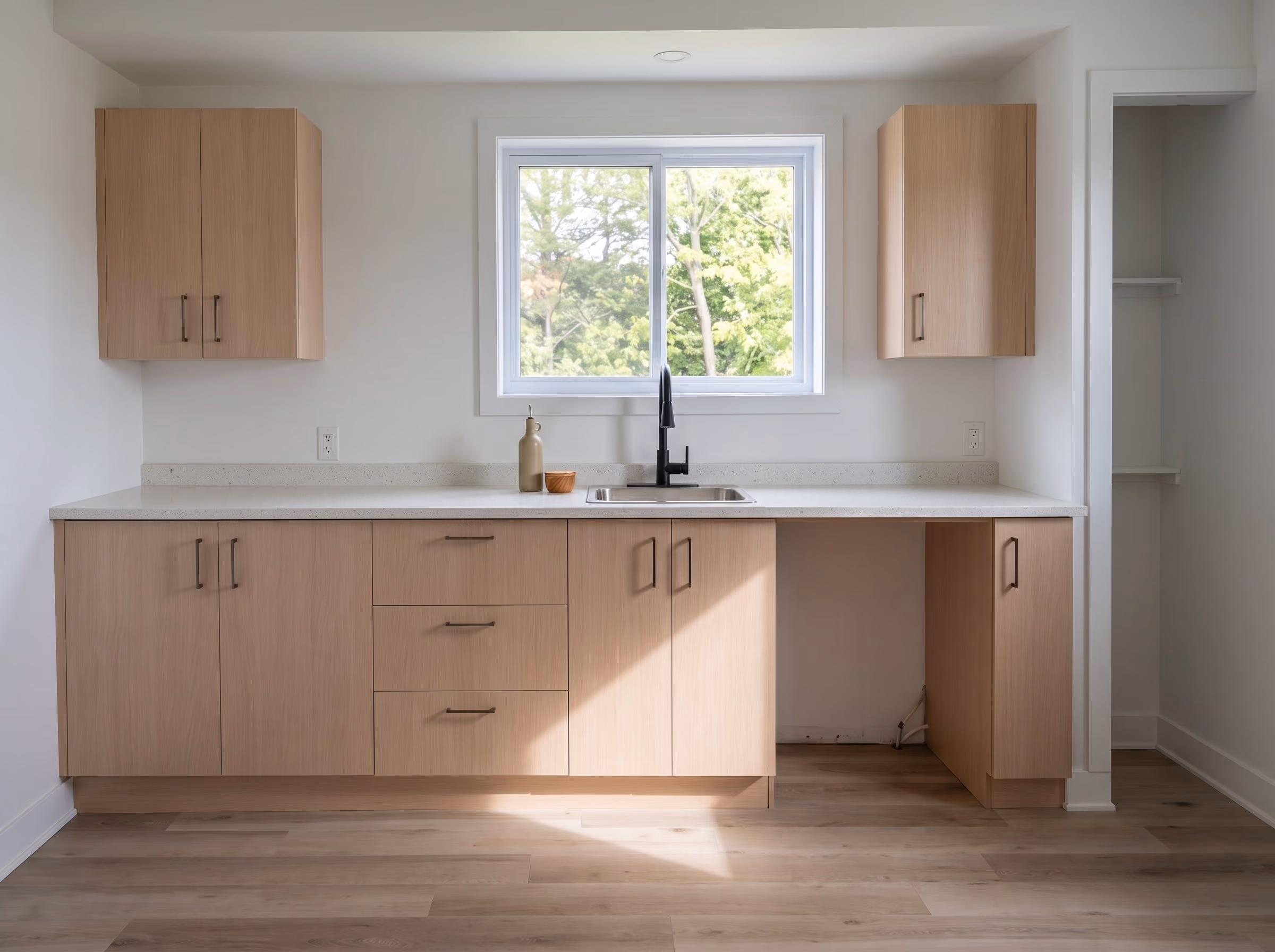 Bright kitchen with pale wood cabinets, slider window above the sink, garden trees sharp through the glass.