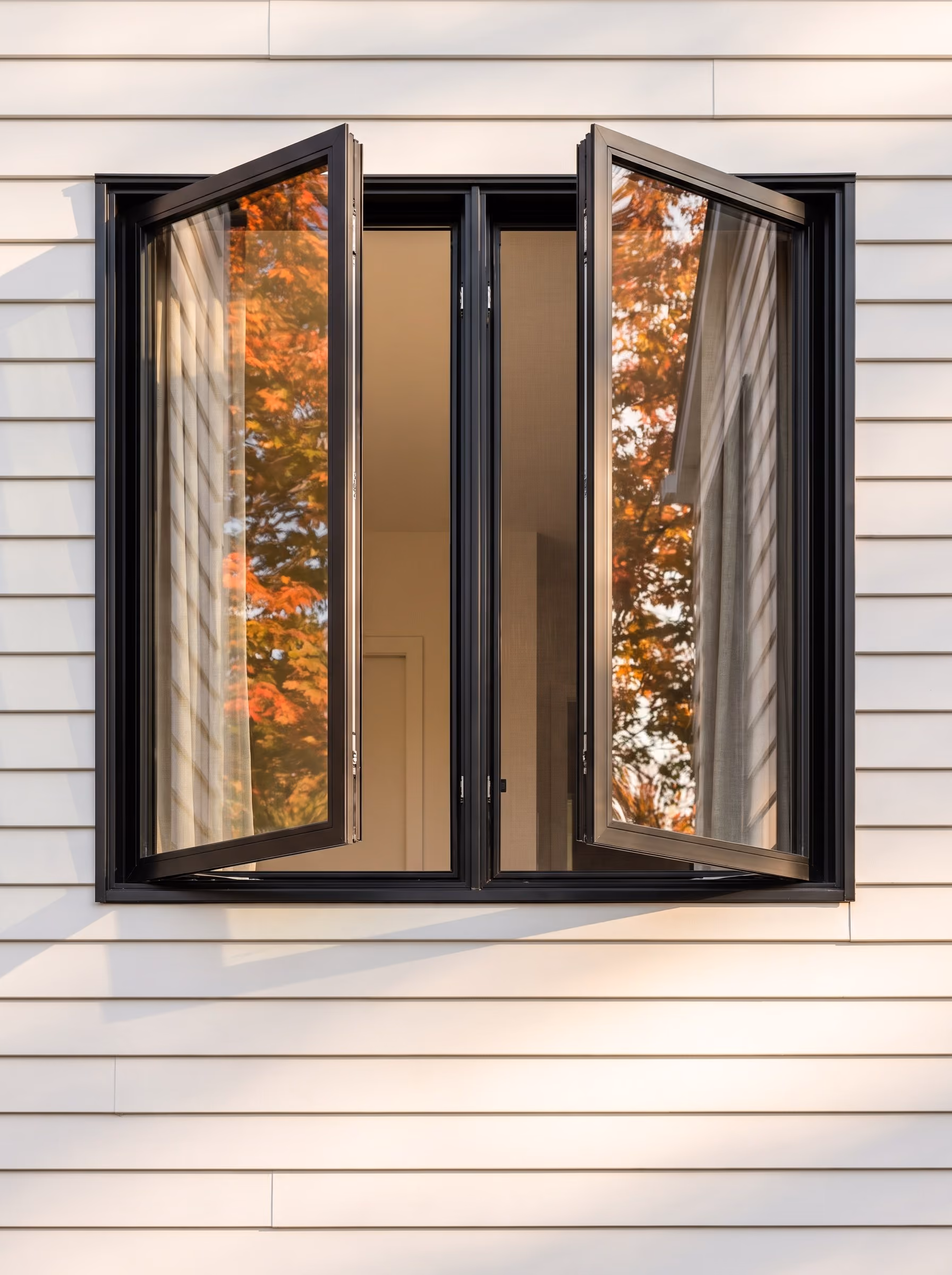 Black-framed double casement window, both sashes open to 90 degrees against white siding, an autumn maple reflected in the glass.
